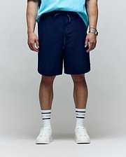 A person stands facing forward in a light blue shirt, Psycho Bunny MENS BIG AND TALL SAMOS HYDROCHROMIC SWIM TRUNK (B9W453F200), white sneakers, white socks with black stripes, and a wristwatch. The background is plain and neutral; face not visible.