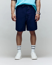 A person stands facing forward in a light blue shirt, Psycho Bunny MENS BIG AND TALL SAMOS HYDROCHROMIC SWIM TRUNK (B9W453F200), white sneakers, white socks with black stripes, and a wristwatch. The background is plain and neutral; face not visible.