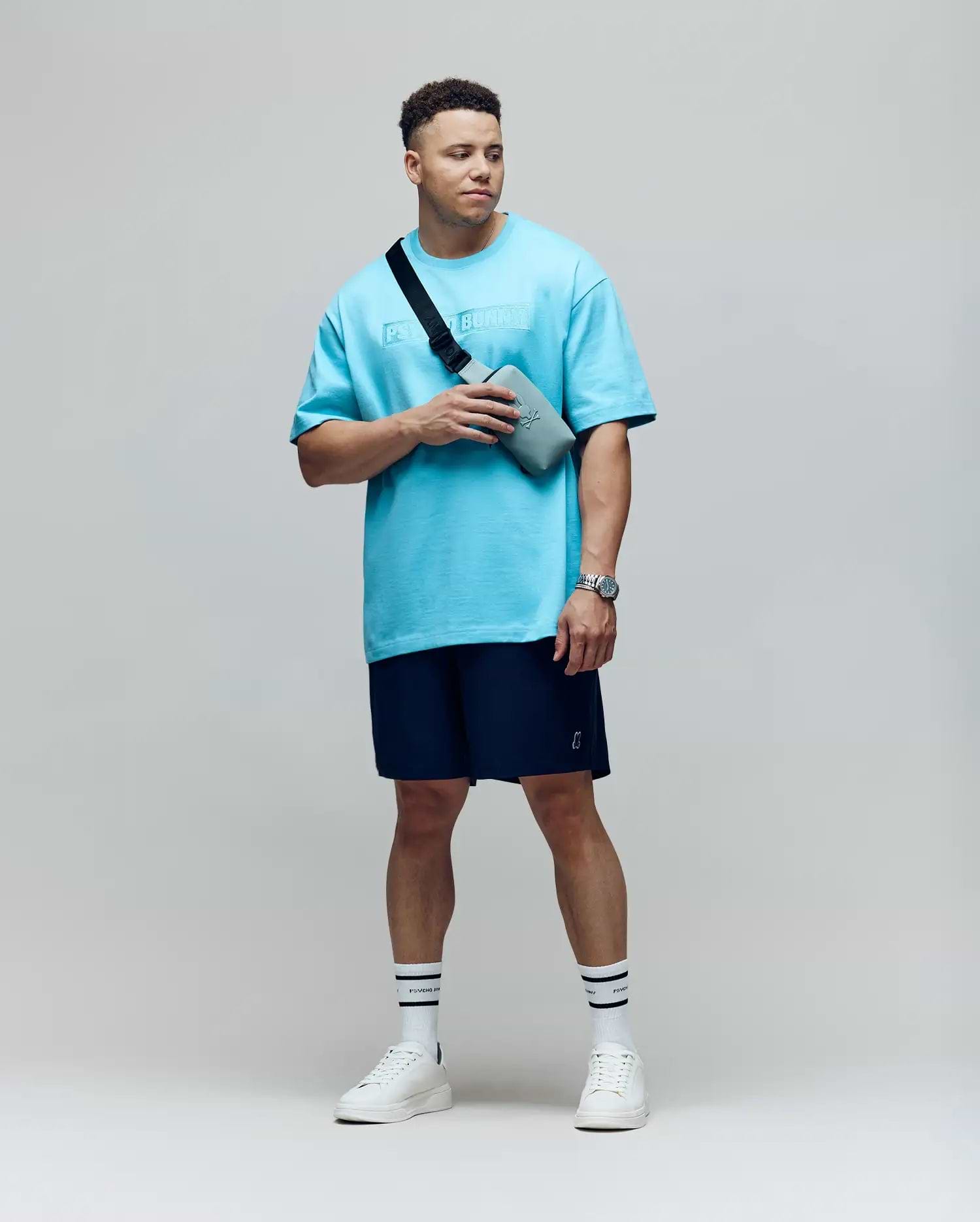 A man stands against a plain background wearing Psycho Bunny MENS BIG AND TALL SAMOS HYDROCHROMIC SWIM TRUNK (B9W453F200), a light blue oversized tee, white sneakers, striped socks, a watch, and a gray crossbody bag, looking to the side.