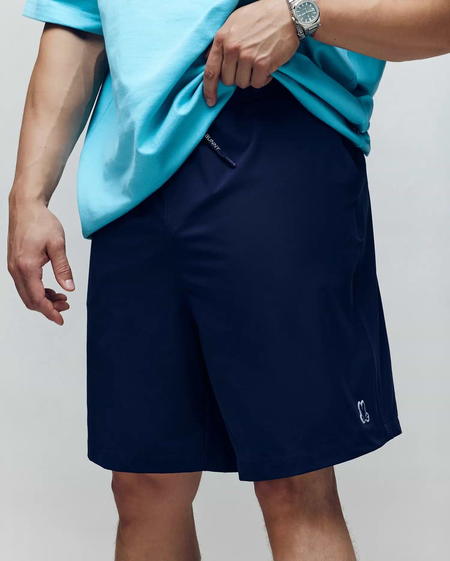A person wearing Psycho Bunny’s MENS BIG AND TALL SAMOS HYDROCHROMIC SWIM TRUNK (B9W453F200), featuring a small embroidered logo near the hem, a mesh lining, and paired with a turquoise t-shirt and watch.