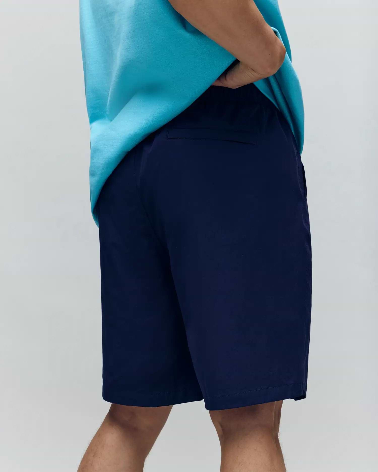 A person wears a loose turquoise t-shirt and Psycho Bunny MENS BIG AND TALL SAMOS HYDROCHROMIC SWIM TRUNK (B9W453F200), standing with one hand in their pocket against a plain light background.