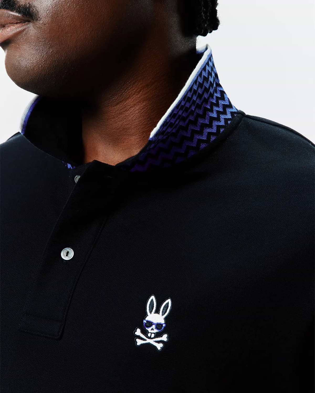 Black Big And Tall Sunny Pique Polo with a white and purple zig-zag patterned inner collar, featuring a small embroidered white Psycho Bunny skull logo with blue sunglasses on the left chest.