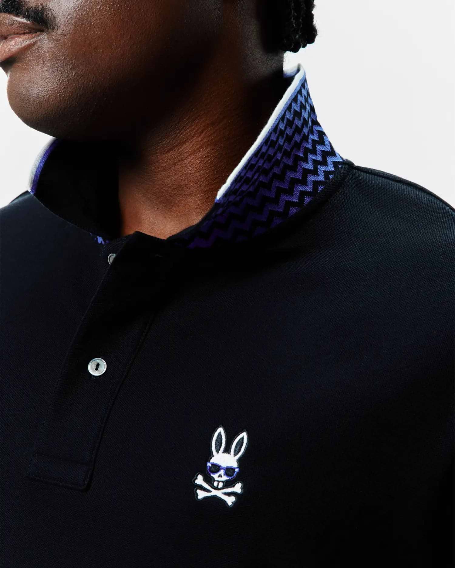 Black Big And Tall Sunny Pique Polo with a white and purple zig-zag patterned inner collar, featuring a small embroidered white Psycho Bunny skull logo with blue sunglasses on the left chest.