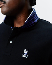 Black Big And Tall Sunny Pique Polo with a white and purple zig-zag patterned inner collar, featuring a small embroidered white Psycho Bunny skull logo with blue sunglasses on the left chest.