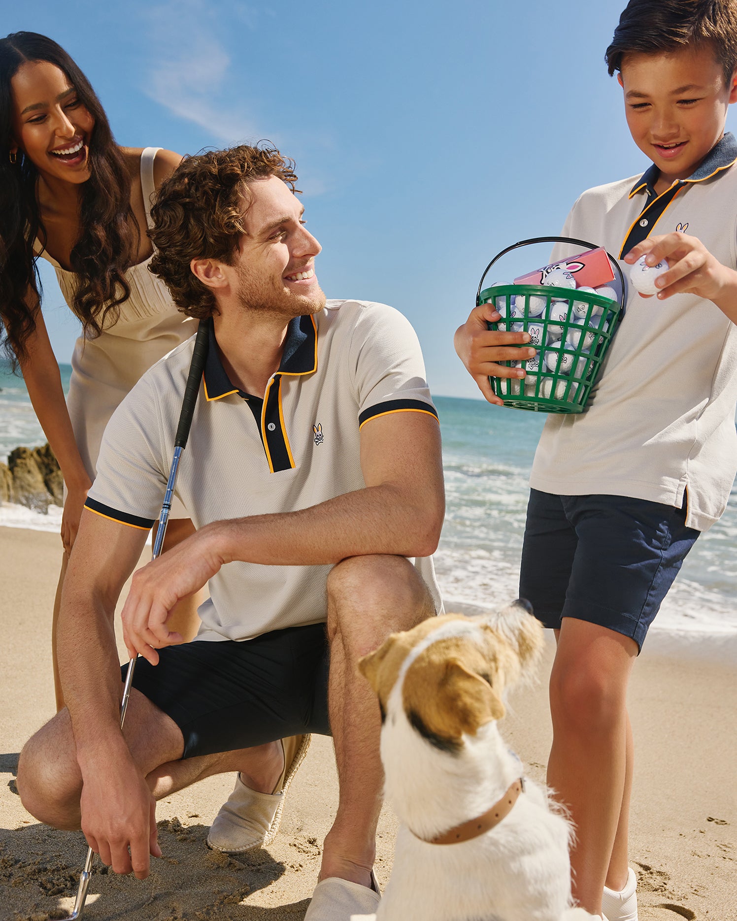 A joyful family on a sunny beach with a young boy handing a green basket to his father, a woman smiling at them, and a small dog looking up, all wearing matching Psycho Bunny MEN'S DALLAS PIQUE POLO SHIRTS.