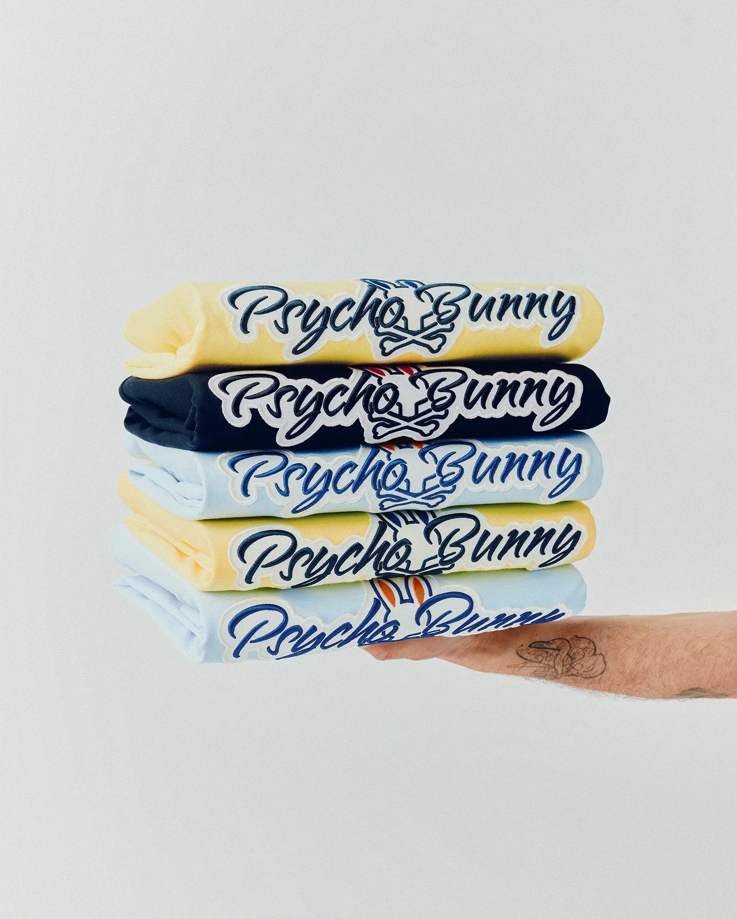 Mens Jaydon Wordmark Graphic Tee stack: yellow, navy, and light blue jersey tees featuring a large embroidered dark blue Psycho Bunny wordmark with white outline and iconic bunny skull logo with red accents on the chest.