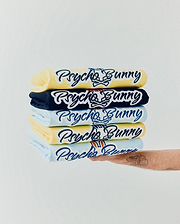 Mens Jaydon Wordmark Graphic Tee stack: light yellow, navy, and light blue jersey tees with large, embroidered Psycho Bunny script logo and iconic bunny skull, some featuring red/orange ear accents.
