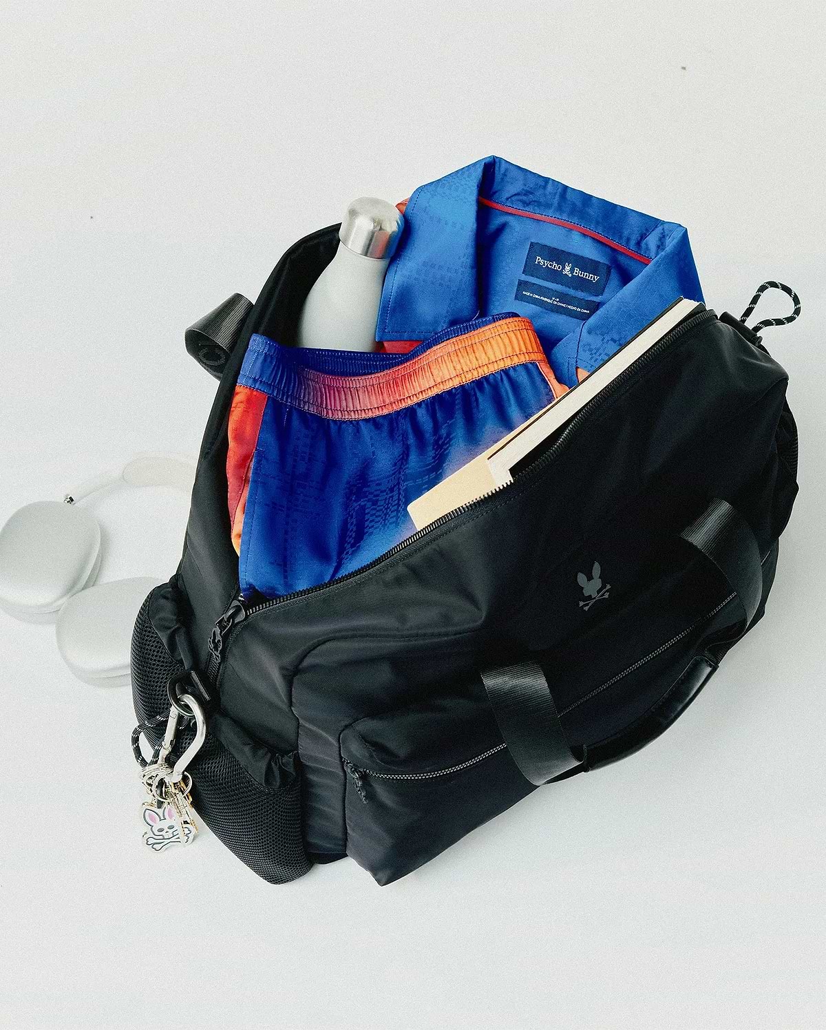 Black Mens Nylon Duffle Bag with a subtle Psycho Bunny skull logo, containing a blue patterned polo with red collar trim and brand tag, plus blue and orange patterned shorts. (6)