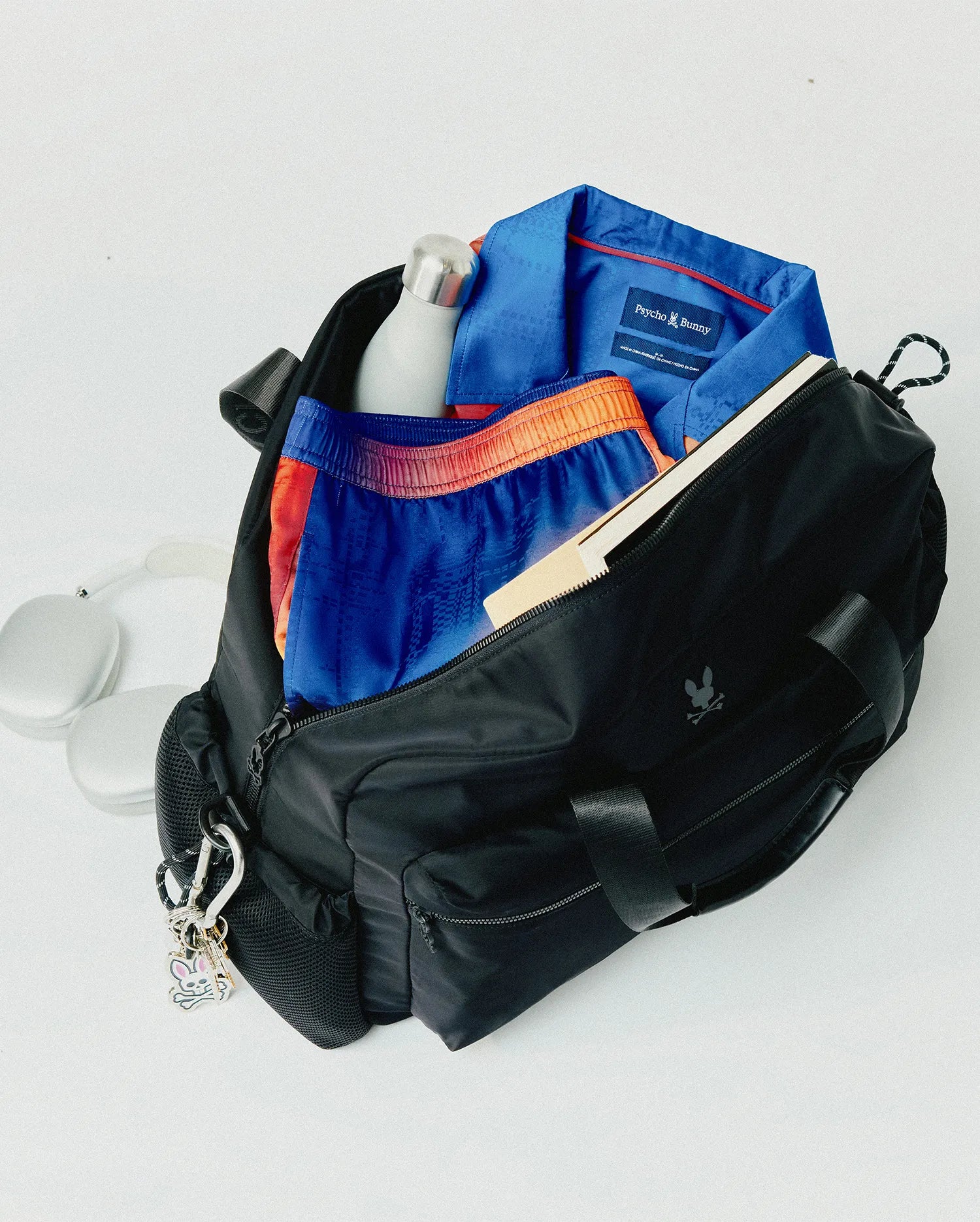 Black Mens Nylon Duffle Bag with a subtle Psycho Bunny skull logo, containing a blue patterned polo with red collar trim and brand tag, plus blue and orange patterned shorts. (6)