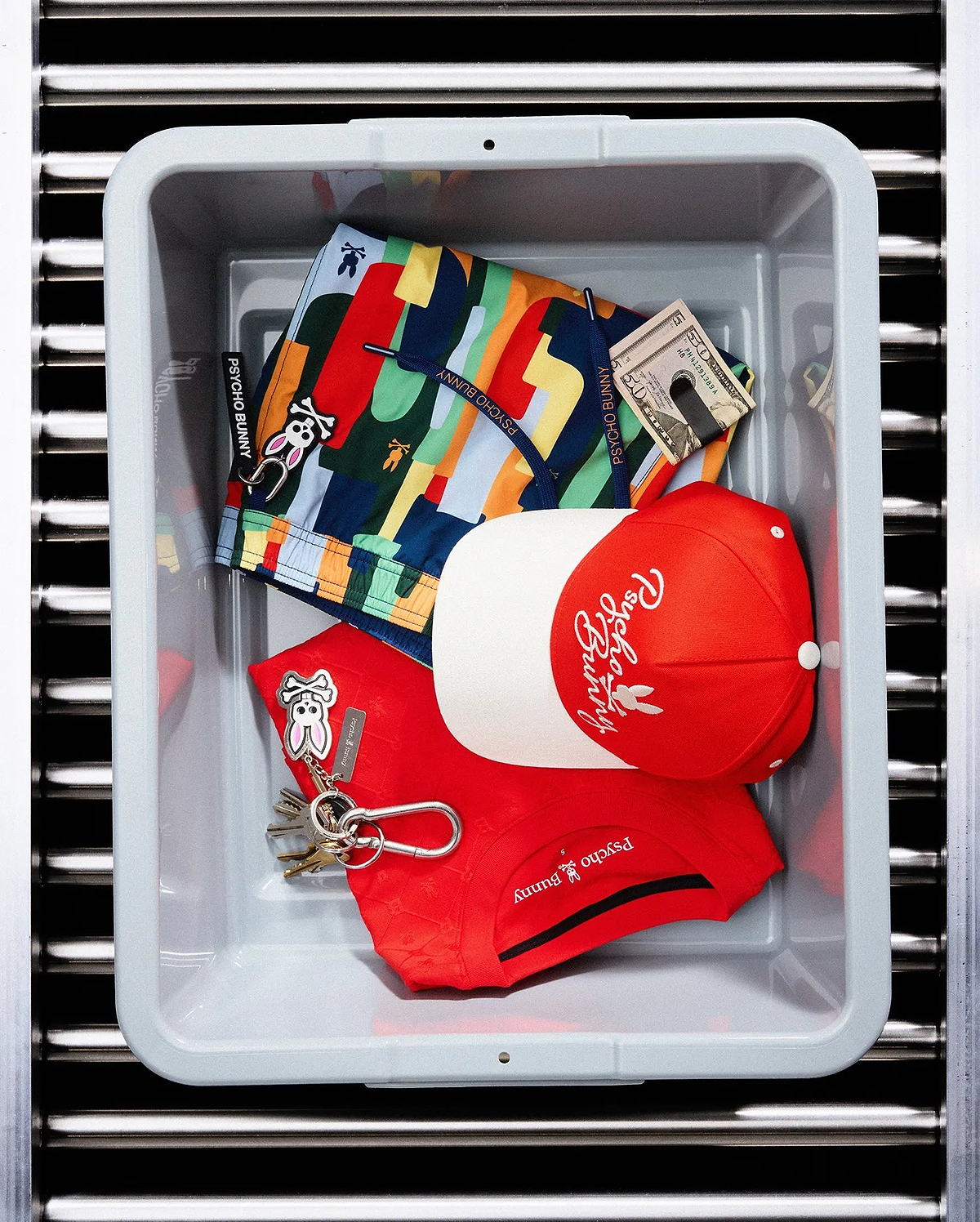 Red pique polo with chest Psycho Bunny logo, multi-color patterned swim shorts featuring bunny skull print and keychain, and a red and white Mens Sable A-Frame Trucker Cap with embroidered Psycho Bunny script.