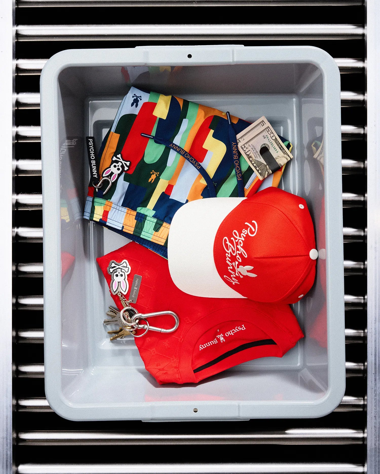 Red pique polo with chest Psycho Bunny logo, multi-color patterned swim shorts featuring bunny skull print and keychain, and a red and white Mens Sable A-Frame Trucker Cap with embroidered Psycho Bunny script.