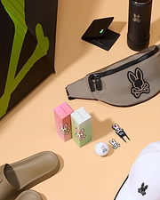 An assortment of various items on a beige surface, including a tan fanny pack, two boxes with Psycho Bunny logos, a golf ball with a bunny logo, BALL MARKER - B6A698C200 from Psycho Bunny, a black wallet, a black bottle, tan slide sandals, a white hat with the Psycho Bunny logo, and a black umbrella from the golfers collection.