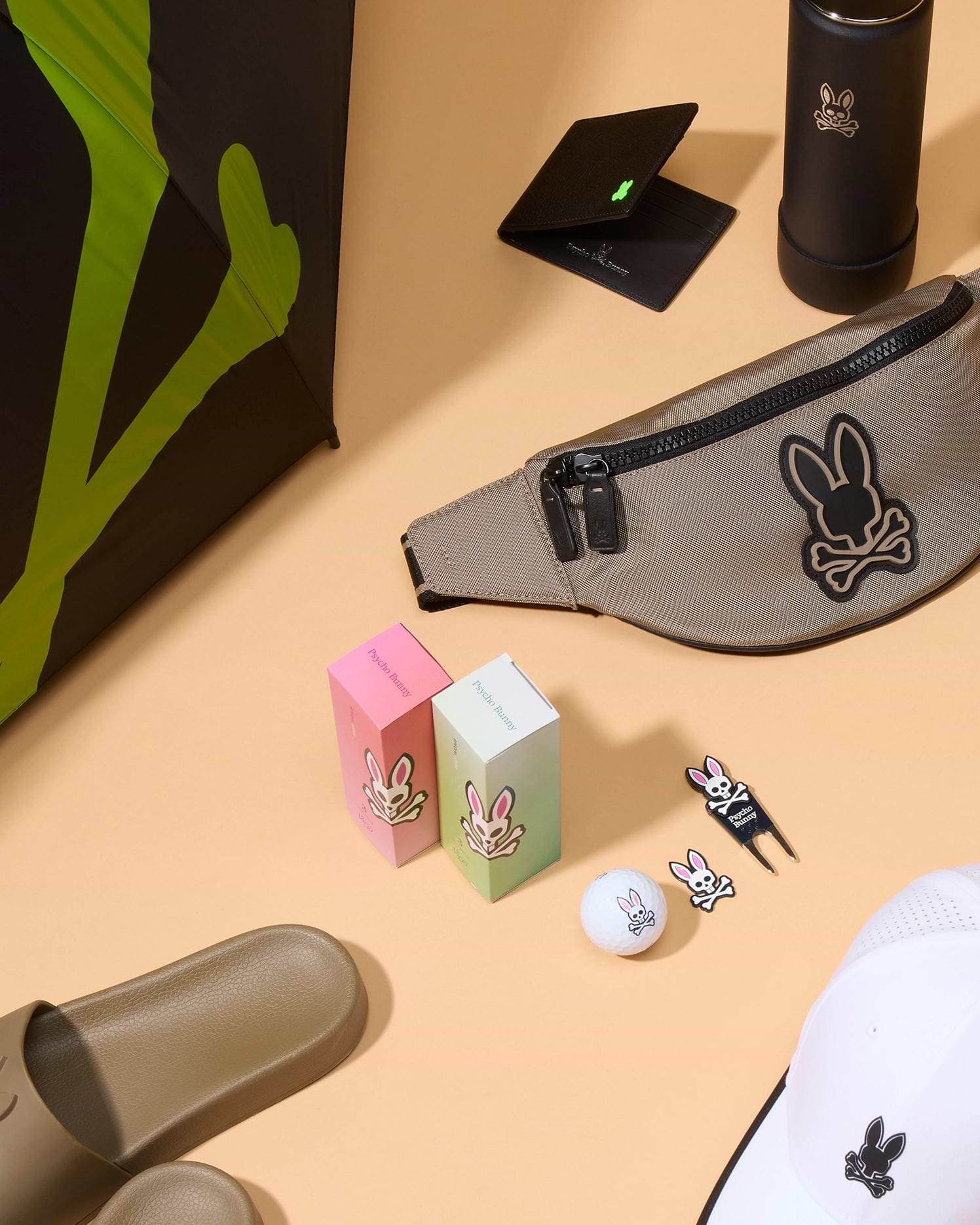 An assortment of various items on a beige surface, including a tan fanny pack, two boxes with Psycho Bunny logos, a golf ball with a bunny logo, BALL MARKER - B6A698C200 from Psycho Bunny, a black wallet, a black bottle, tan slide sandals, a white hat with the Psycho Bunny logo, and a black umbrella from the golfers collection.