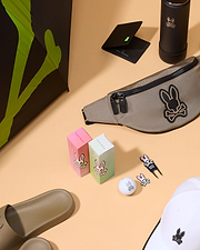 Various golf-related accessories are arranged on a beige surface. Items include a golf umbrella, a waist bag with a bunny skull logo, golf balls and tees, a cap, two boxes of golf gifts featuring a magnetic ball marker and Psycho Bunny DIVOT TOOL - B6A699C200, a water bottle, a black wallet, and a single slide sandal.