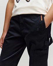 A person wears Psycho Bunny KIDS RUSSELL CARPENTER PANT - KB0600001 in black, featuring an embroidered bunny logo, cargo pockets, and an orange label by the pocket; a white graphic shirt is partially visible. One hand rests in the pocket.