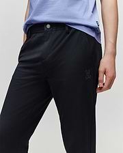 A person wears the Psycho Bunny KIDS MATHIEU COMMUTER PANT (KB0600002) featuring a small Bunny and crossbones logo on the pocket, paired with a light purple shirt, resting their hand on their thigh.