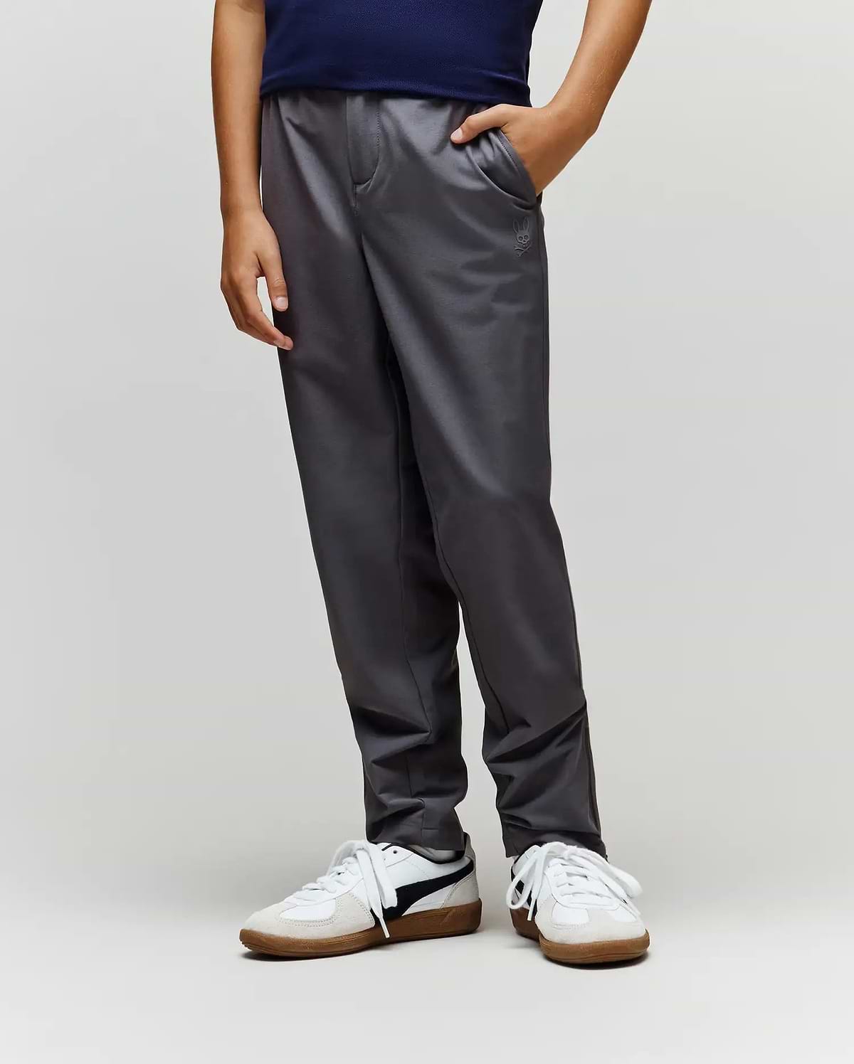 A child stands with one hand in their pocket, wearing Psycho Bunny's KIDS MATHIEU COMMUTER PANT (KB0600002) in dark gray, featuring subtle Bunny embroidery, paired with a navy shirt and white sneakers against a light background.