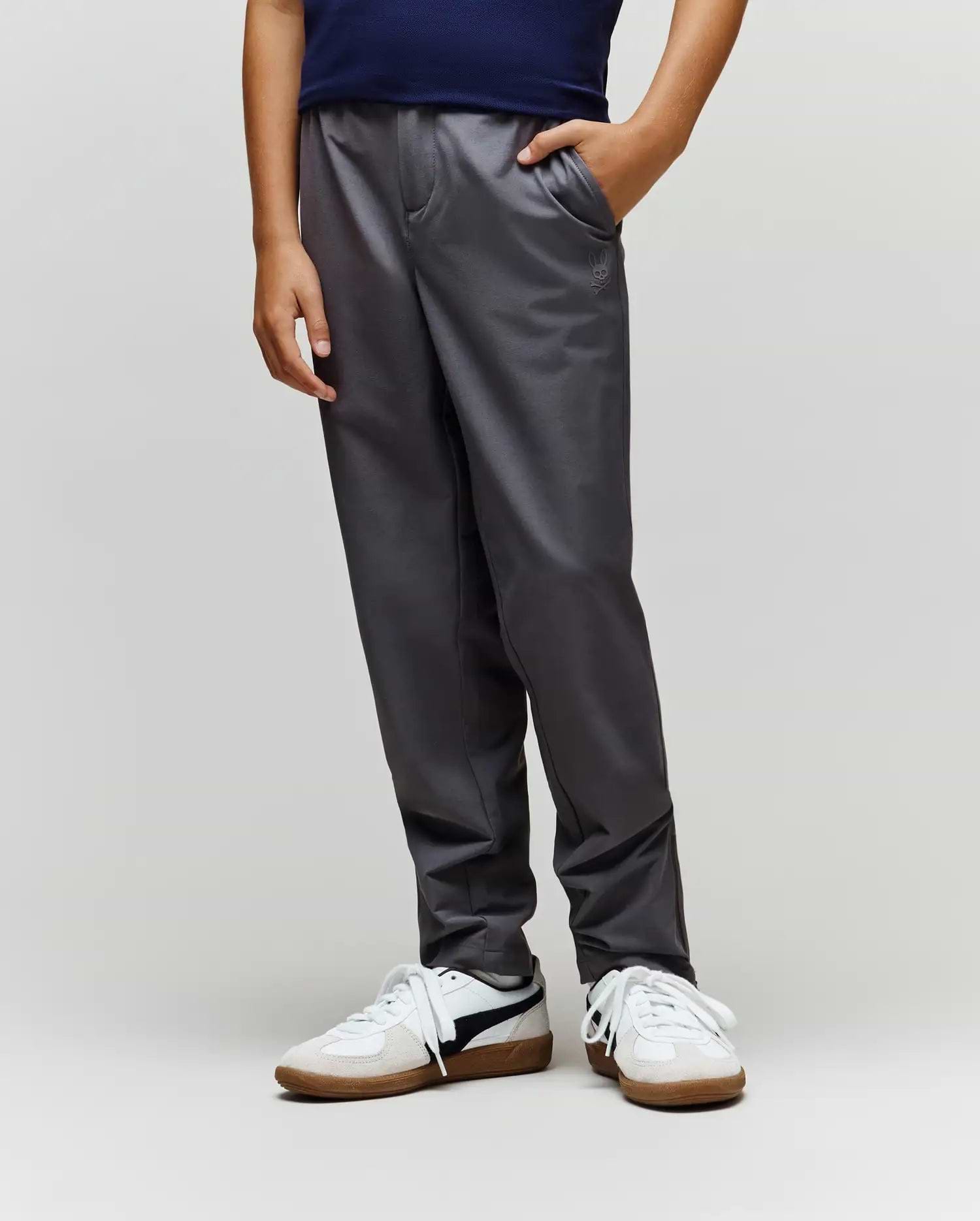 A child stands with one hand in their pocket, wearing Psycho Bunny's KIDS MATHIEU COMMUTER PANT (KB0600002) in dark gray, featuring subtle Bunny embroidery, paired with a navy shirt and white sneakers against a light background.