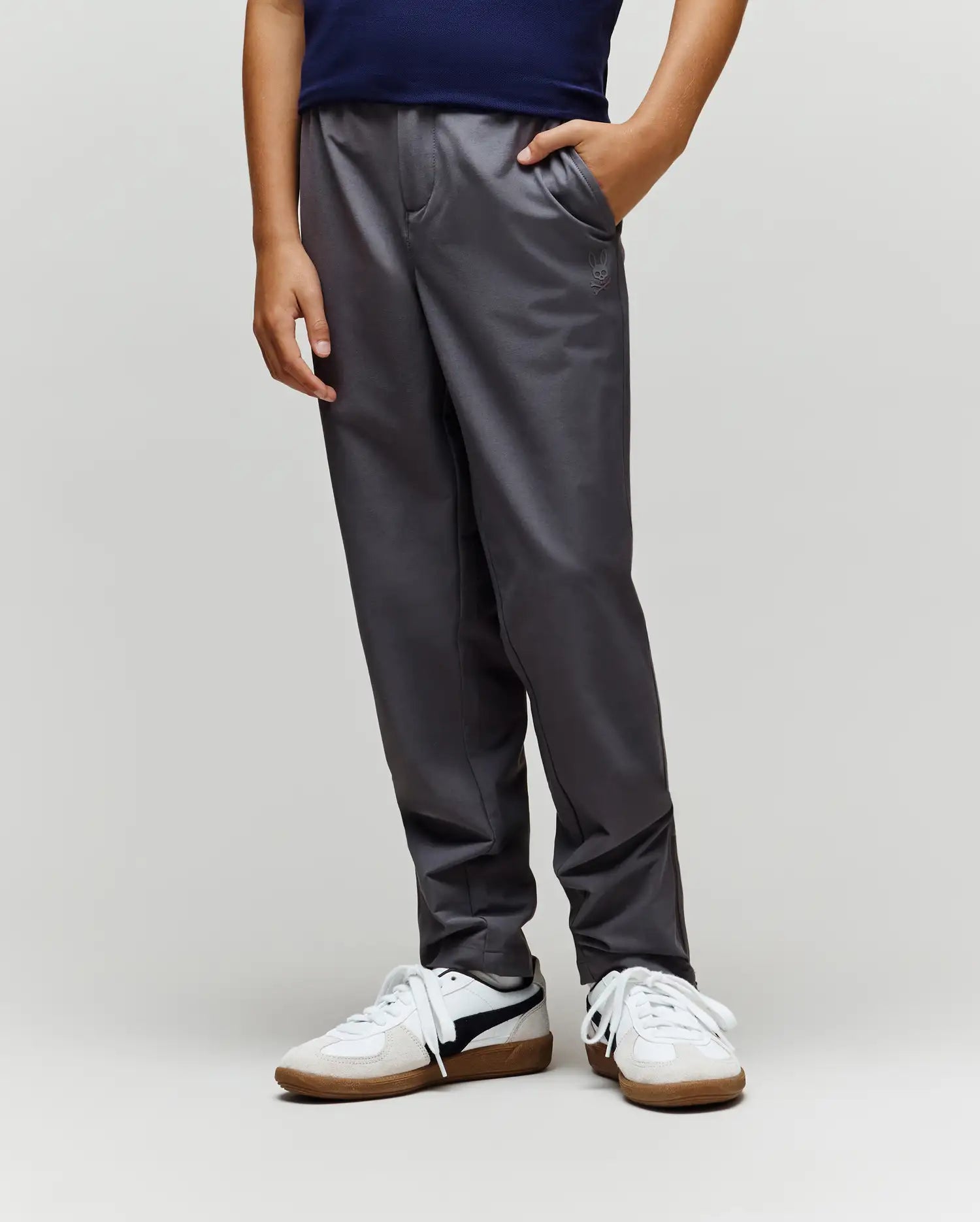 A child stands with one hand in their pocket, wearing Psycho Bunny's KIDS MATHIEU COMMUTER PANT (KB0600002) in dark gray, featuring subtle Bunny embroidery, paired with a navy shirt and white sneakers against a light background.