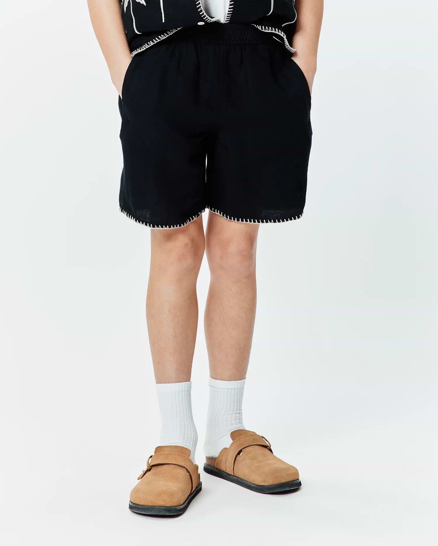 Kids Brad Linen Short, black relaxed fit linen shorts with elastic waistband, white contrast stitching on hem, and a subtle embroidered Psycho Bunny skull logo on the lower left leg.
