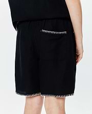 Kids Brad Linen Short in black, a relaxed fit garment with visible linen texture, accented by distinctive white blanket stitching on the back patch pocket and along the bottom hem.