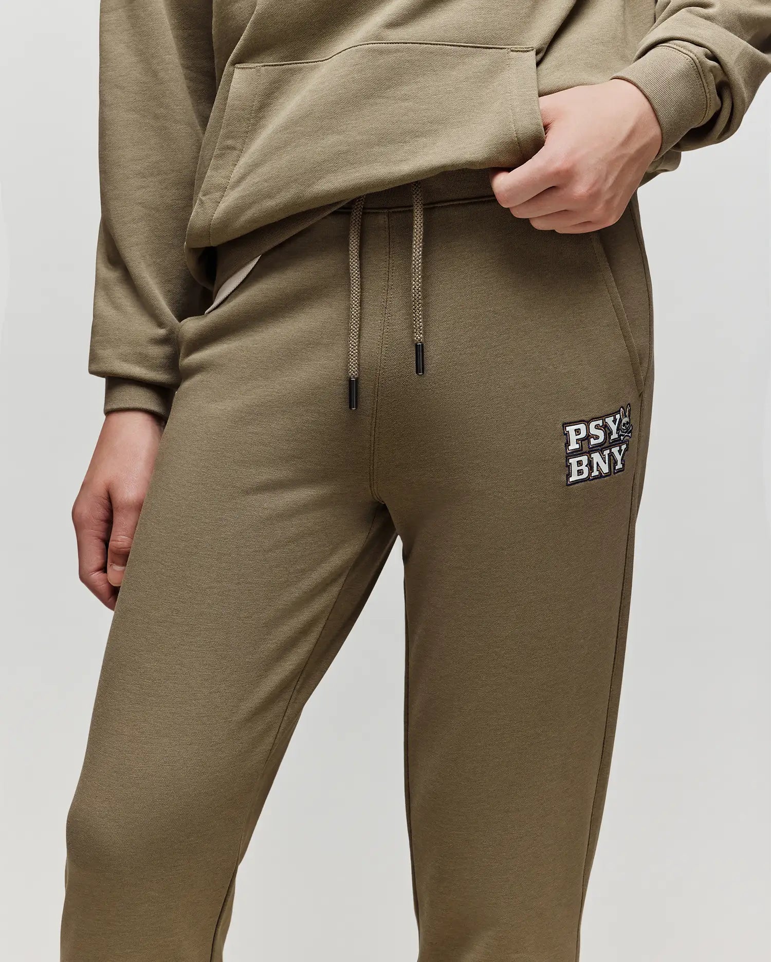 A person wears the Psycho Bunny KIDS WADE SWEATPANT (KB0800002) in olive green, featuring "PSY BNYY" and a graphic on the left thigh, paired with a matching sweatshirt. Made from a premium cotton blend, one hand is in the pocket, the other on the hip.