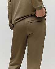 A child wears the Psycho Bunny KIDS WADE SWEATPANT (KB0800002) in olive green, premium cotton blend, posing from behind with one hand on the waistband near a welt pocket, set against a plain light background.