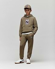 A young boy faces forward against a plain background, wearing Psycho Bunny’s KIDS WADE SWEATPANT (KB0800002) in tan with jogger pants, a hoodie featuring the cartoon bunny logo, a matching cap, and white sneakers. His hands are in his pockets.