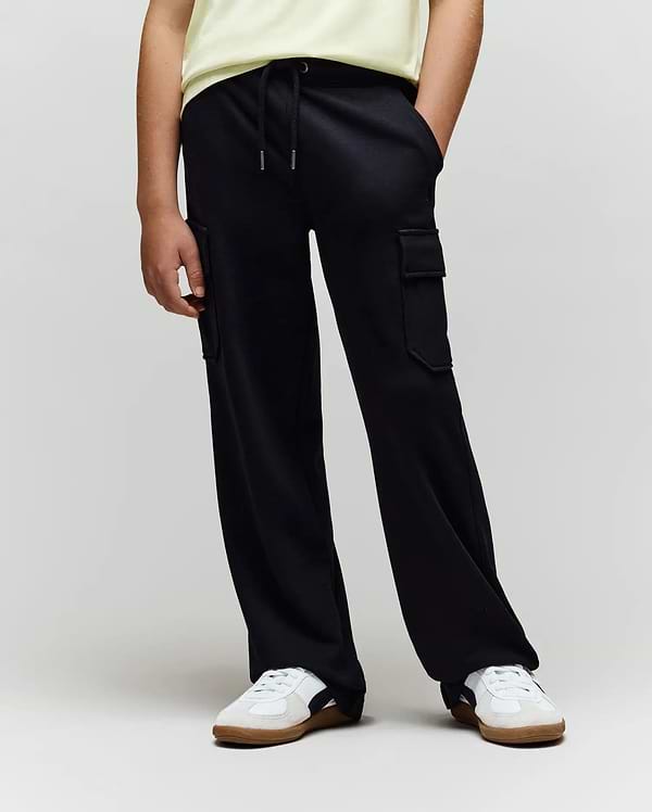 A person in a light yellow shirt, white sneakers with beige soles, and loose black Psycho Bunny KIDS NASH CARGO SWEATPANT (KB0800004) with cargo pockets stands against a plain background with hands in their pockets.