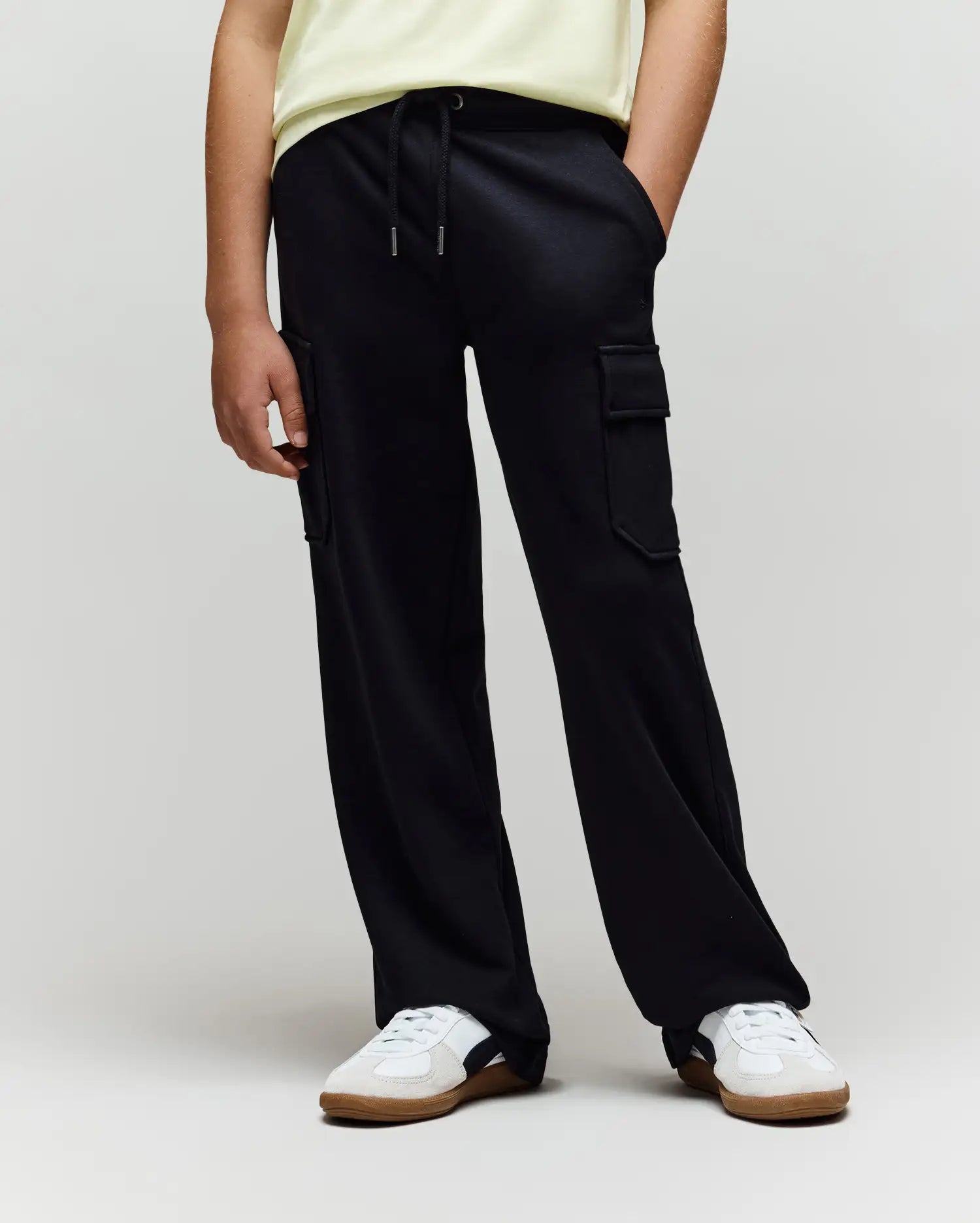 A person in a light yellow shirt, white sneakers with beige soles, and loose black Psycho Bunny KIDS NASH CARGO SWEATPANT (KB0800004) with cargo pockets stands against a plain background with hands in their pockets.