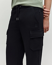 A person wearing Psycho Bunny KIDS NASH CARGO SWEATPANT - KB0800004—black cargo jogger sweatpants with large pockets, a drawstring waist, subtle stitching, and a small embroidered logo near the pocket—shown from the waist down.