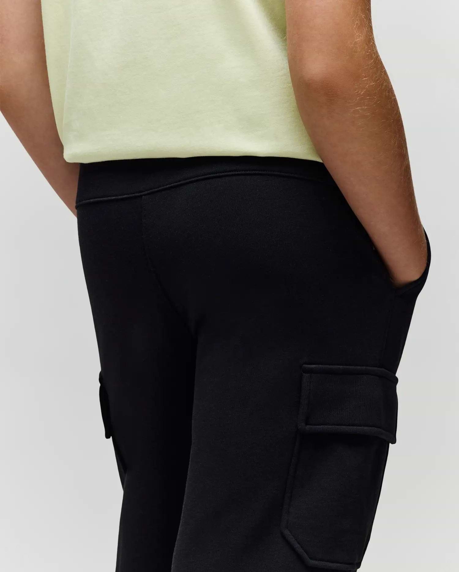 A child wears Psycho Bunny's KIDS NASH CARGO SWEATPANT (KB0800004) in black with cargo pockets and a light yellow shirt, hands in pockets; image cropped from waist to mid-thigh.