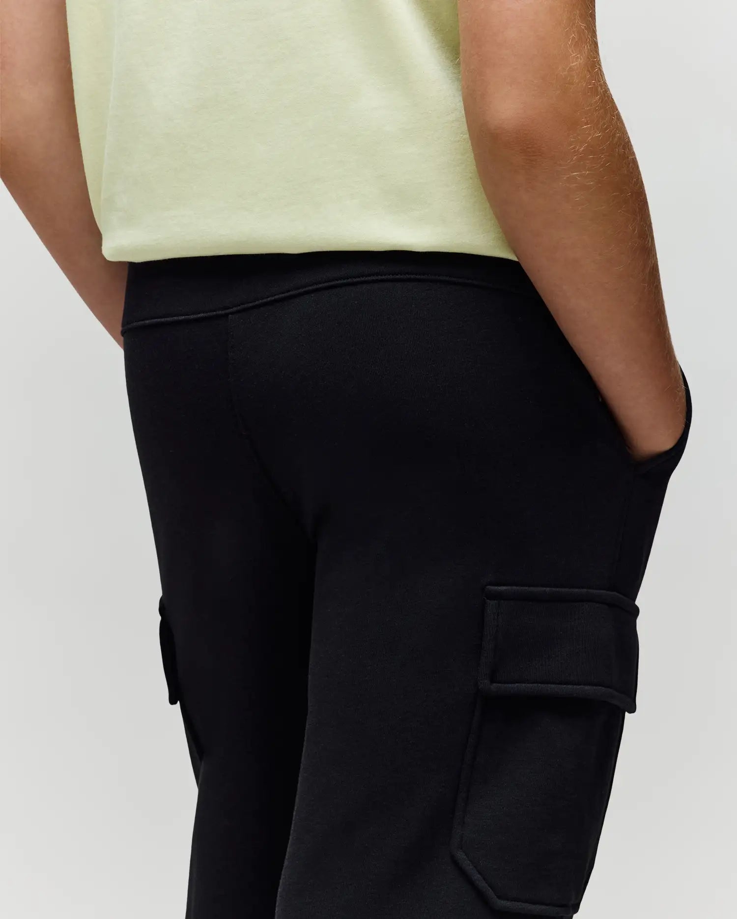 A child wears Psycho Bunny's KIDS NASH CARGO SWEATPANT (KB0800004) in black with cargo pockets and a light yellow shirt, hands in pockets; image cropped from waist to mid-thigh.