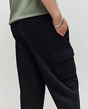 A person wears a light green shirt and Psycho Bunny KIDS NASH CARGO SWEATPANT - KB0800004, showing off its spacious cargo pockets with one hand in the pocket and a silver bracelet on their wrist, set against a plain background.