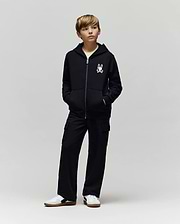 A young boy stands with hands in his pockets, wearing a black zip-up hoodie with Psycho Bunny’s cartoon bunny logo, KIDS NASH CARGO SWEATPANT (KB0800004) with cargo pockets, and white sneakers against a plain light gray background.