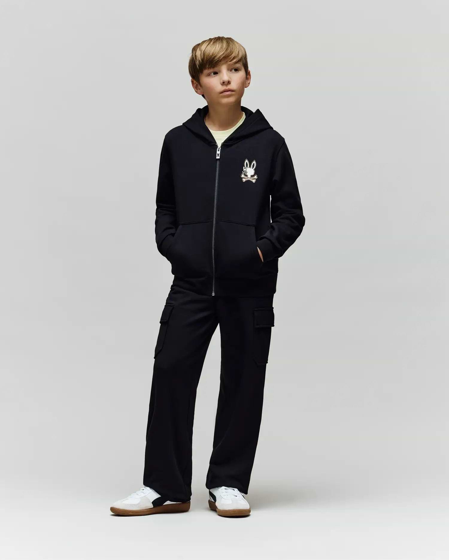 A young boy stands with hands in his pockets, wearing a black zip-up hoodie with Psycho Bunny’s cartoon bunny logo, KIDS NASH CARGO SWEATPANT (KB0800004) with cargo pockets, and white sneakers against a plain light gray background.