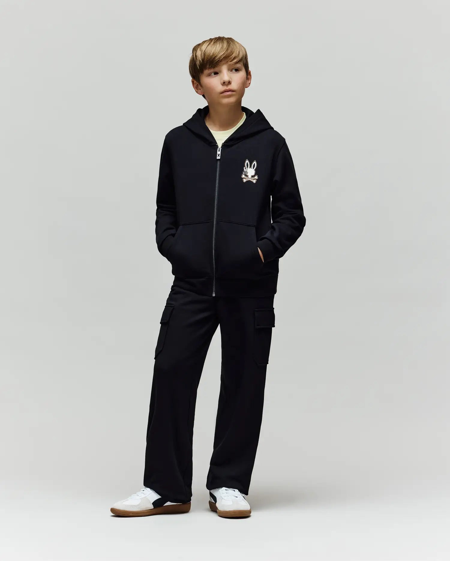A young boy stands with hands in his pockets, wearing a black zip-up hoodie with Psycho Bunny’s cartoon bunny logo, KIDS NASH CARGO SWEATPANT (KB0800004) with cargo pockets, and white sneakers against a plain light gray background.