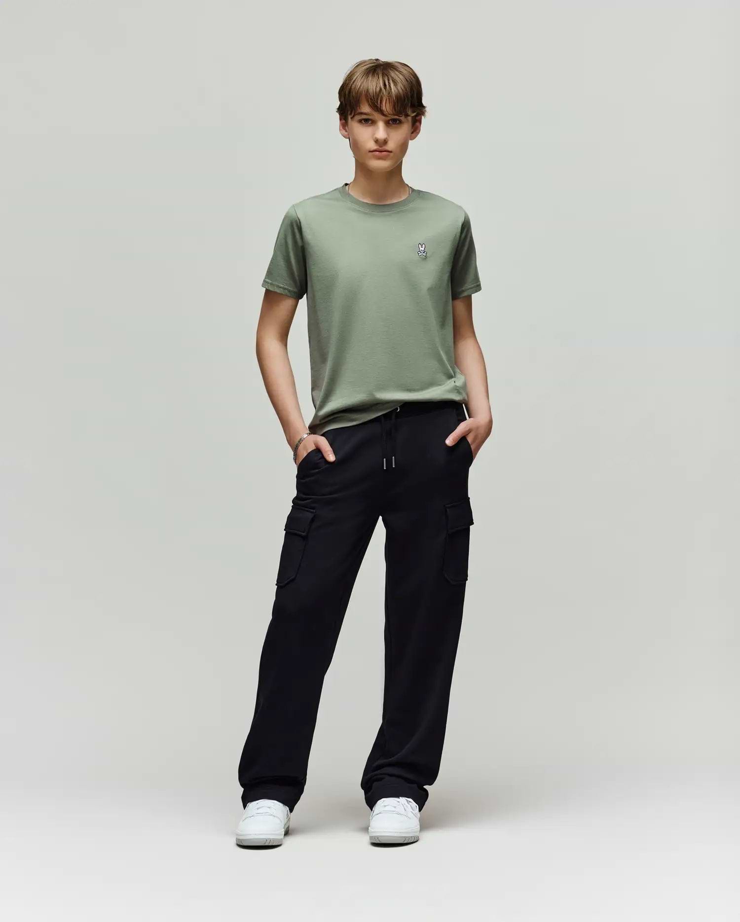 A young person stands in a studio against a light background, wearing Psycho Bunny KIDS NASH CARGO SWEATPANT - KB0800004 with handy cargo pockets, a light green tee, and white sneakers for ultimate sweatpant comfort. Hands are tucked in their pockets.