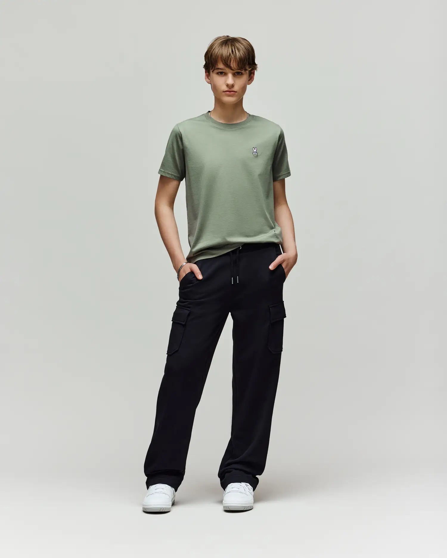 A young person stands in a studio against a light background, wearing Psycho Bunny KIDS NASH CARGO SWEATPANT - KB0800004 with handy cargo pockets, a light green tee, and white sneakers for ultimate sweatpant comfort. Hands are tucked in their pockets.