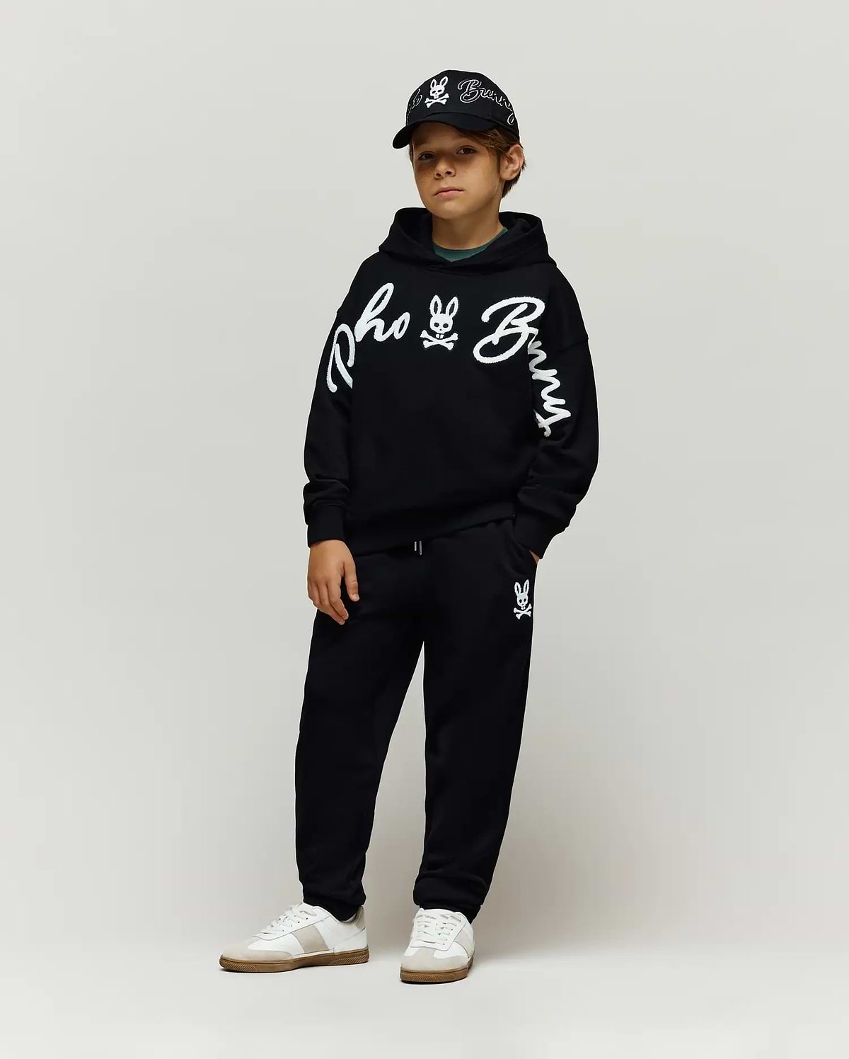 Black relaxed hoodie with large white chenille Psycho Bunny script and sleeve logo, Kids Tate Chenille Sweatpant with small bunny skull logo, and a black cap with white embroidered logos.