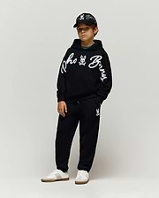 Black relaxed hoodie with large white chenille Psycho Bunny script and sleeve logo, Kids Tate Chenille Sweatpant with small bunny skull logo, and a black cap with white embroidered logos.