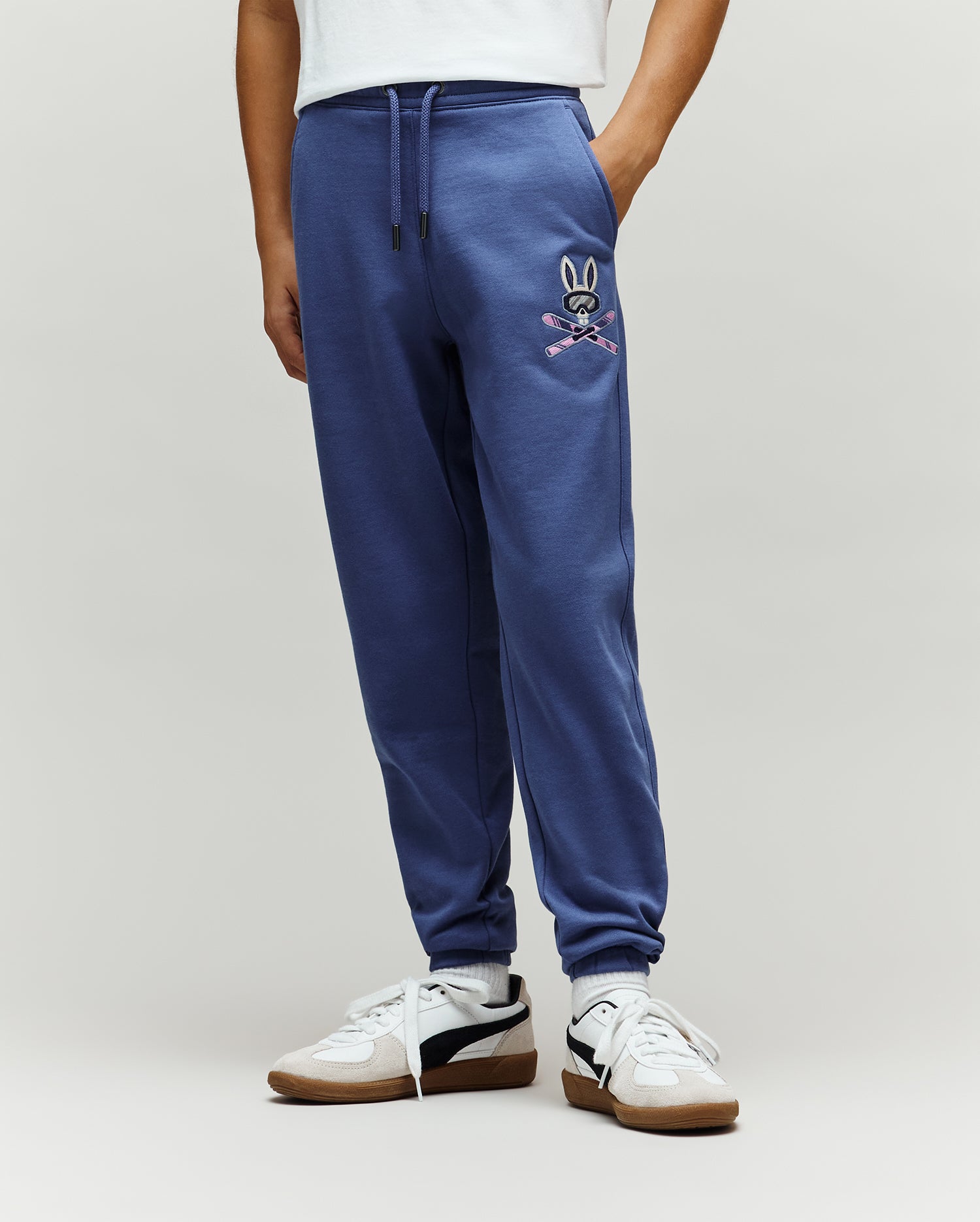 Kids Indigo Ski Bunny Sweatpant, a relaxed fit french terry jogger in deep blue, featuring a vibrant embroidered Psycho Bunny skull with goggles and crossed pink skis on the left thigh.