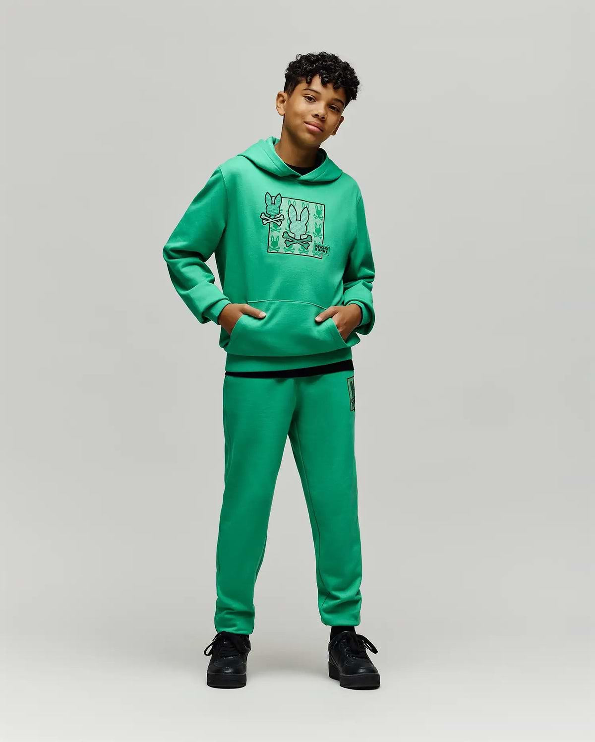 Bright green kids' hoodie with a large Psycho Bunny skull grid graphic on the chest and a kangaroo pocket, matching Kids Varen Jogger pants with a small bunny skull logo on the left thigh.