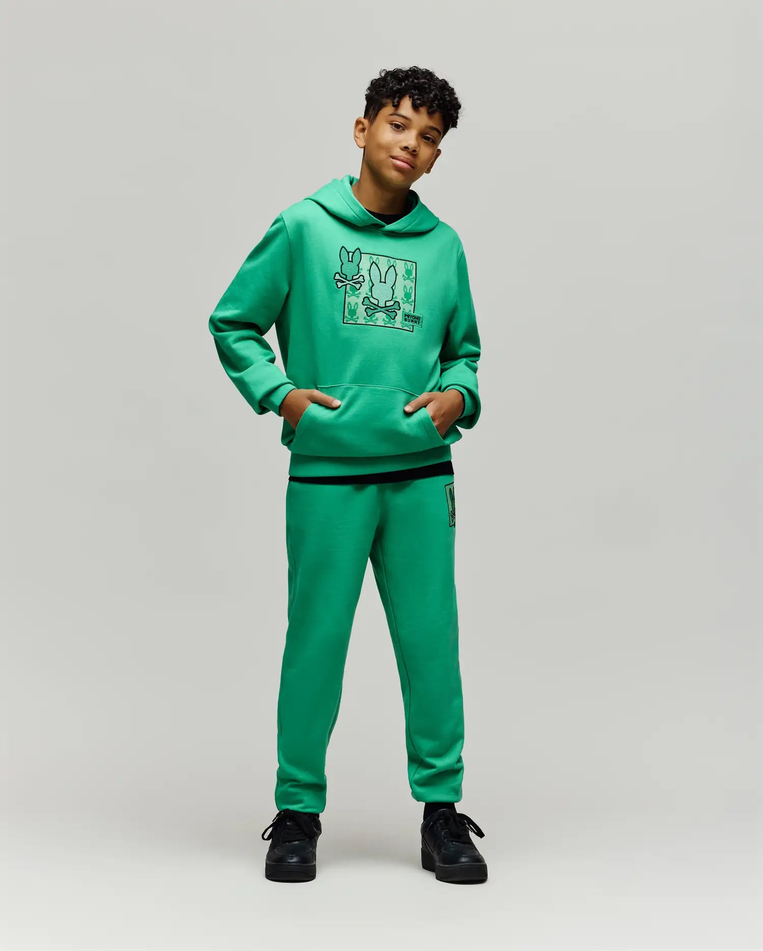 Bright green kids' hoodie with a large Psycho Bunny skull grid graphic on the chest and a kangaroo pocket, matching Kids Varen Jogger pants with a small bunny skull logo on the left thigh.