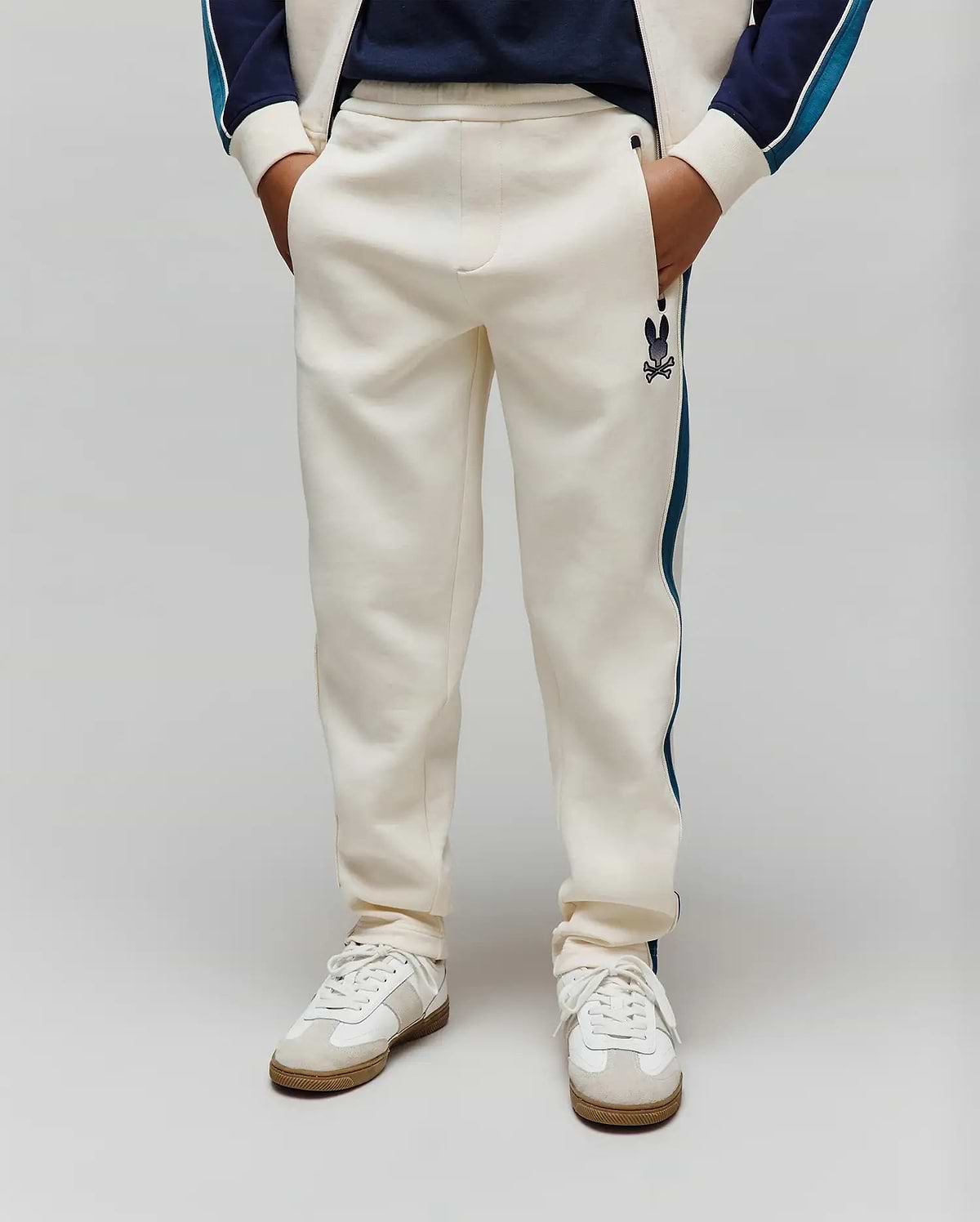 Kids Caspian Contrast Stripe Track Pant in cream french terry, featuring a relaxed fit, dark blue side stripe, and a small embroidered Psycho Bunny skull logo on the left thigh.