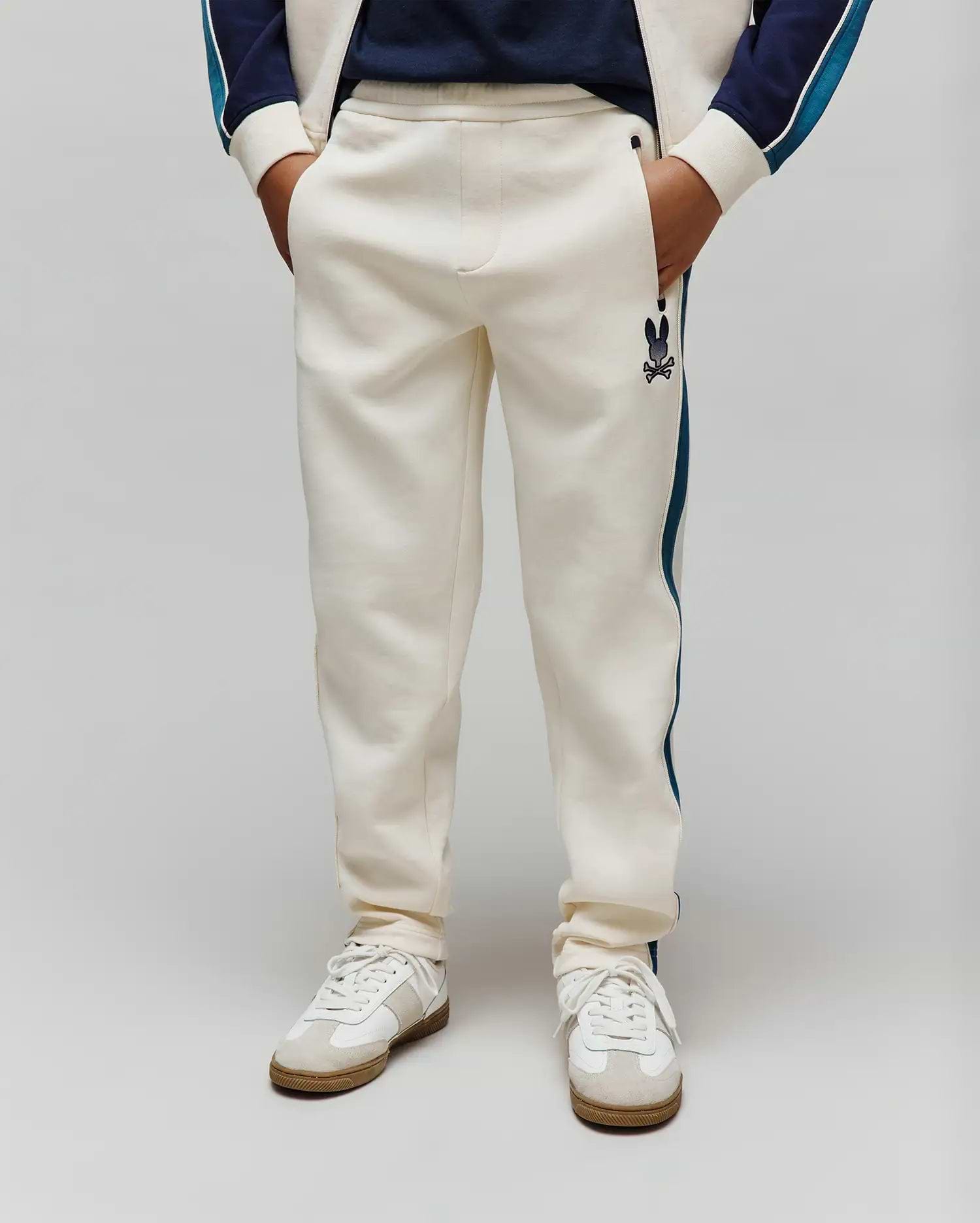Kids Caspian Contrast Stripe Track Pant in cream french terry, featuring a relaxed fit, dark blue side stripe, and a small embroidered Psycho Bunny skull logo on the left thigh.