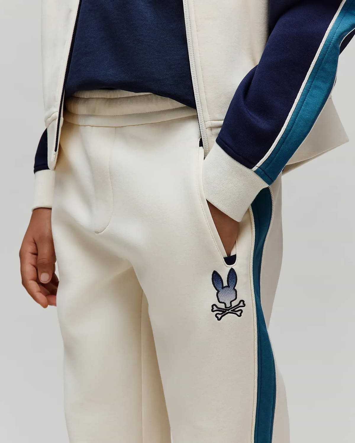 Kids Caspian Contrast Stripe Track Pant in cream french terry, featuring a relaxed fit, navy and teal side stripes with white piping, and an embroidered gradient blue Psycho Bunny skull logo on the left thigh.