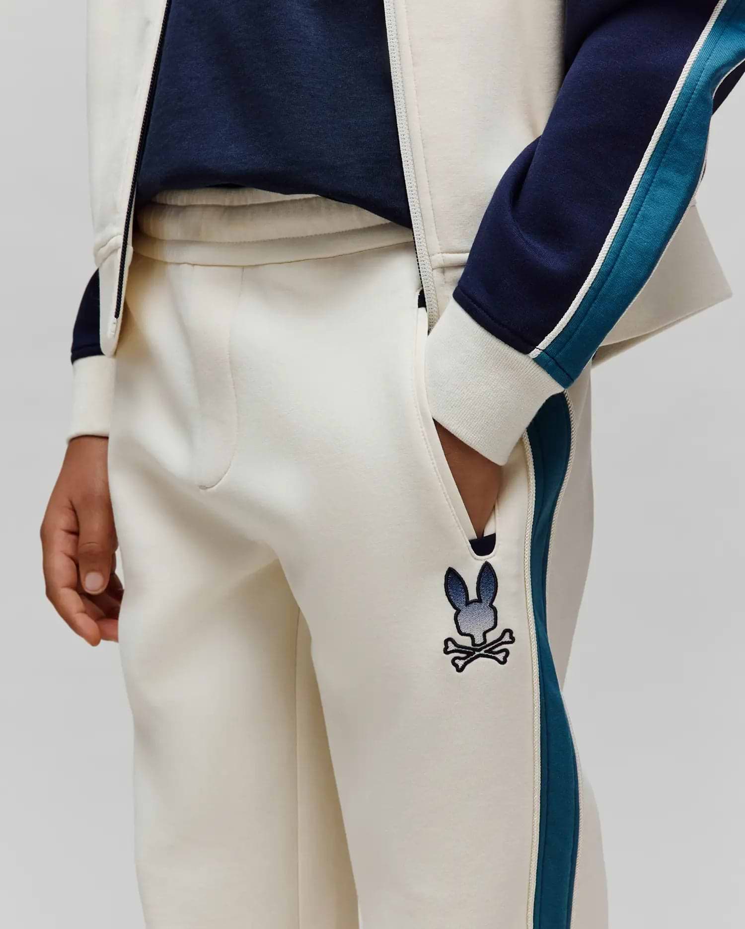 Kids Caspian Contrast Stripe Track Pant in cream french terry, featuring a relaxed fit, navy and teal side stripes with white piping, and an embroidered gradient blue Psycho Bunny skull logo on the left thigh.