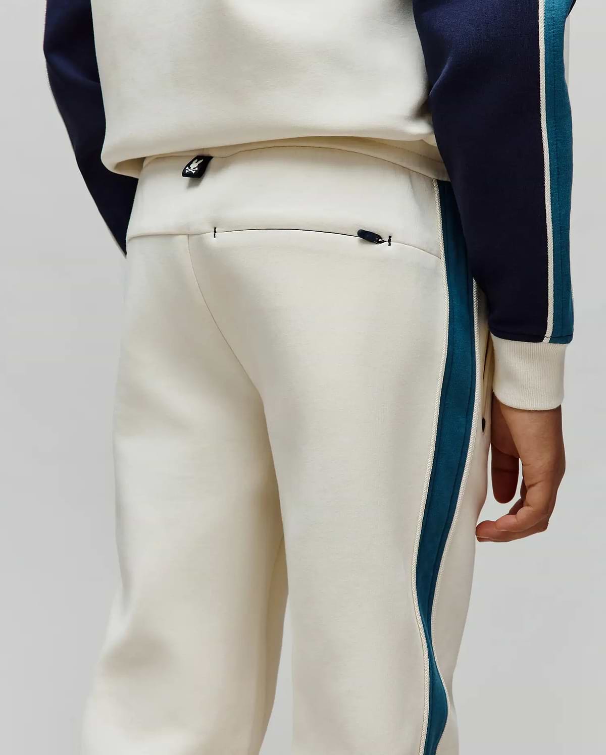 Kids Caspian Contrast Stripe Track Pant in cream french terry, featuring a relaxed fit, teal and white side stripes, a small Psycho Bunny logo tag on the waistband, and a zippered back pocket.