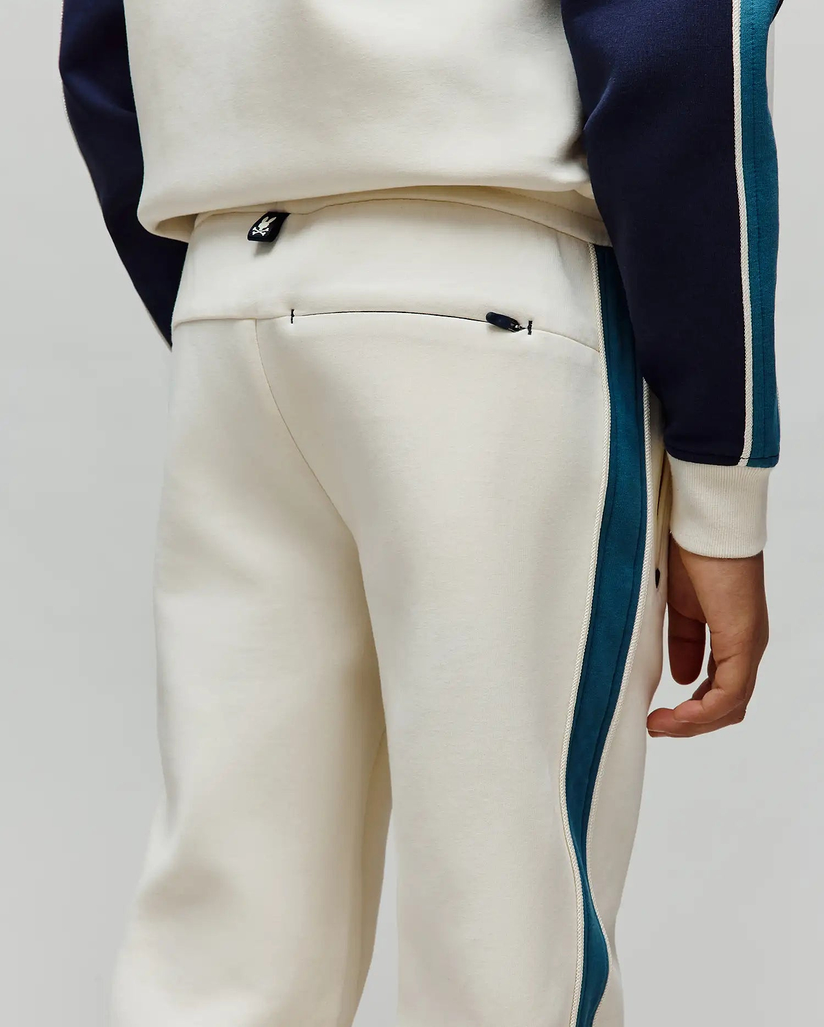 Kids Caspian Contrast Stripe Track Pant in cream french terry, featuring a relaxed fit, teal and white side stripes, a small Psycho Bunny logo tag on the waistband, and a zippered back pocket.